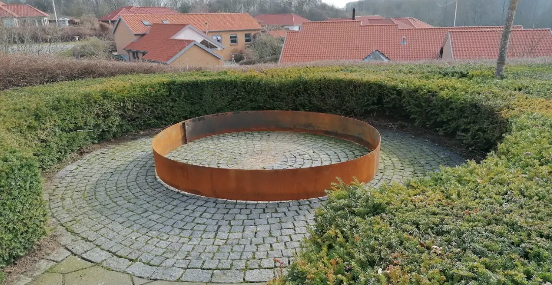 Rund skulptur i metal omgivet af grøn hæk